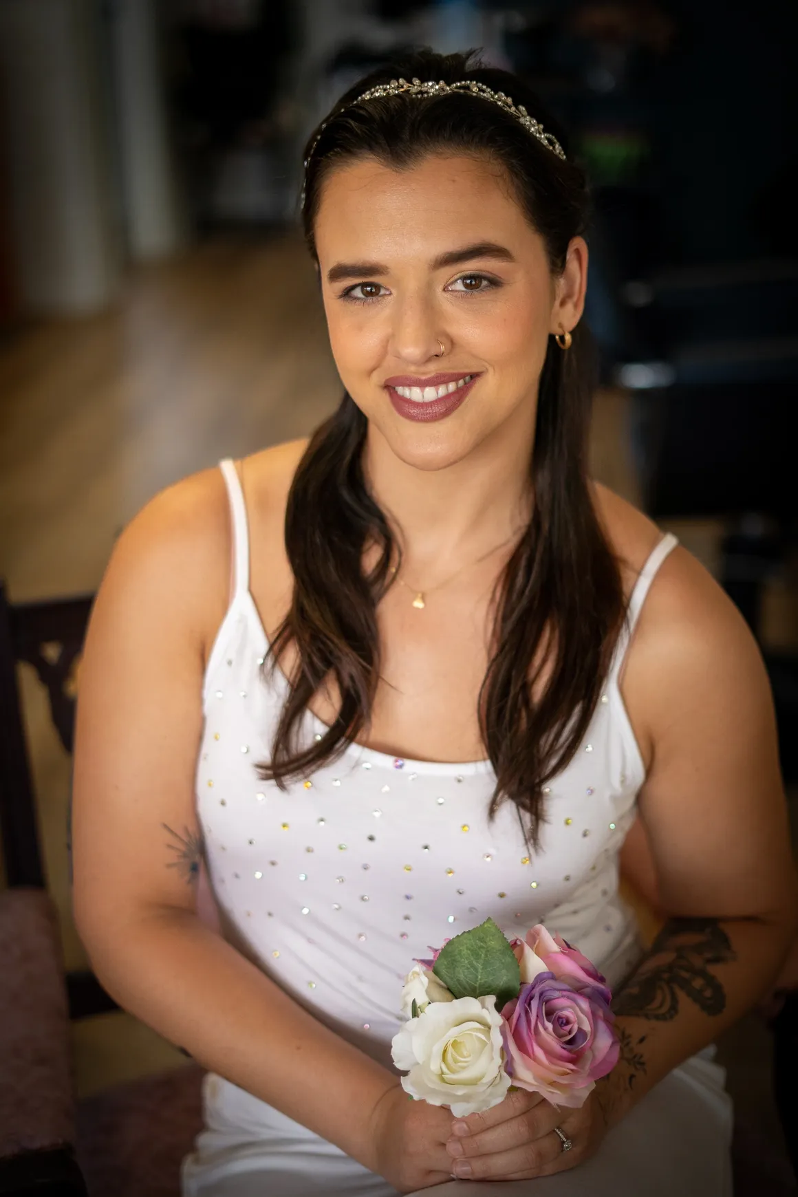 A smiling bride holds a small bouquet of pastel roses while seated indoors. She wears a white, sparkly tank top-style dress and a delicate headband, with her hair styled in soft waves. Her makeup features a warm glow, defined brows, berry-toned lipstick, and subtle shimmer eyeshadow. A tattoo is visible on her forearm, and she wears a nose ring and gold hoop earrings.