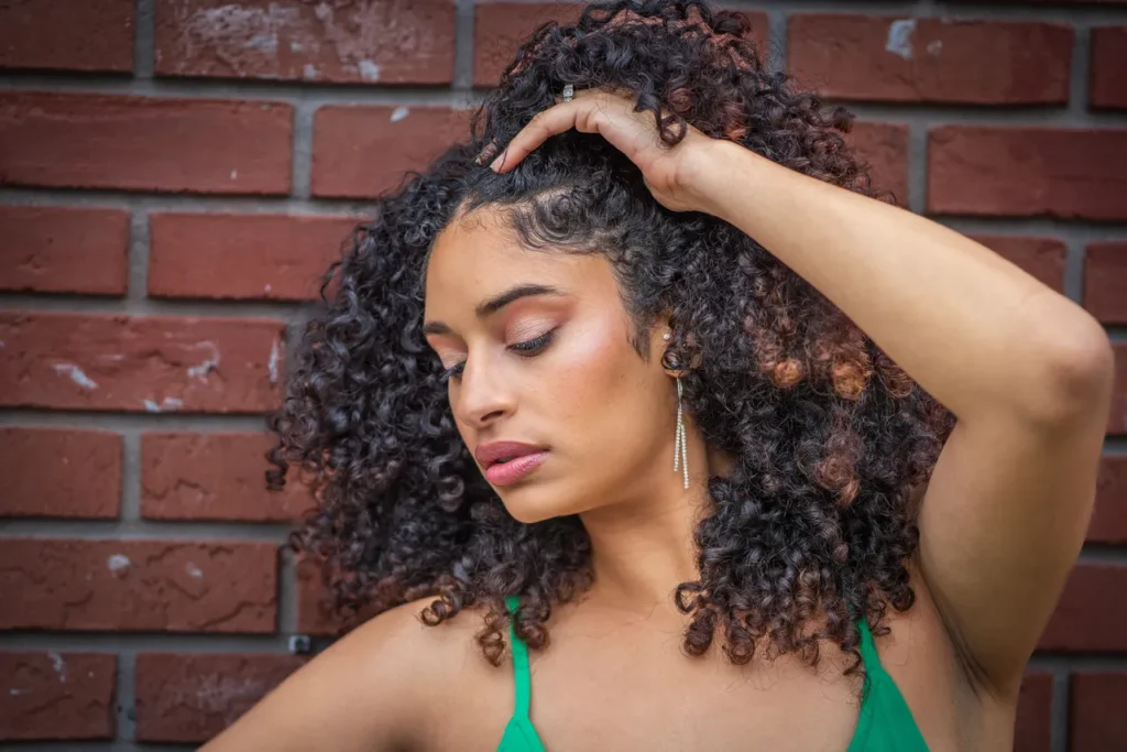 A woman with voluminous, natural curly hair poses in front of a red brick wall, with one hand raised to her head. She wears a green top and long, dangling earrings. Her makeup features a soft rose-toned eyeshadow, defined brows, glowing highlighter, and a mauve lip. Her expression is serene and confident, emphasizing her radiant skin and textured hair.