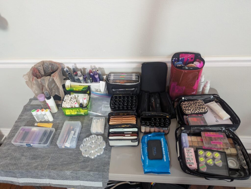 A professional makeup artist’s organized workstation featuring sanitized tools and labeled products. The table is covered with a clean disposable barrier and displays a wide array of makeup essentials, including foundation, concealer, palettes, skincare products, brushes, cotton swabs, and disposables. Multiple pouches and containers hold items neatly, emphasizing hygiene, preparation, and a wide range of shades and formulas suitable for diverse skin tones and needs.