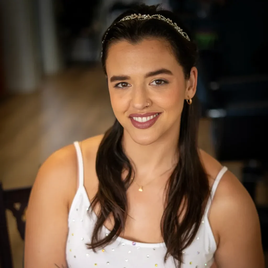 A smiling bride holds a small bouquet of pastel roses while seated indoors. She wears a white, sparkly tank top-style dress and a delicate headband, with her hair styled in soft waves. Her makeup features a warm glow, defined brows, berry-toned lipstick, and subtle shimmer eyeshadow. A tattoo is visible on her forearm, and she wears a nose ring and gold hoop earrings.