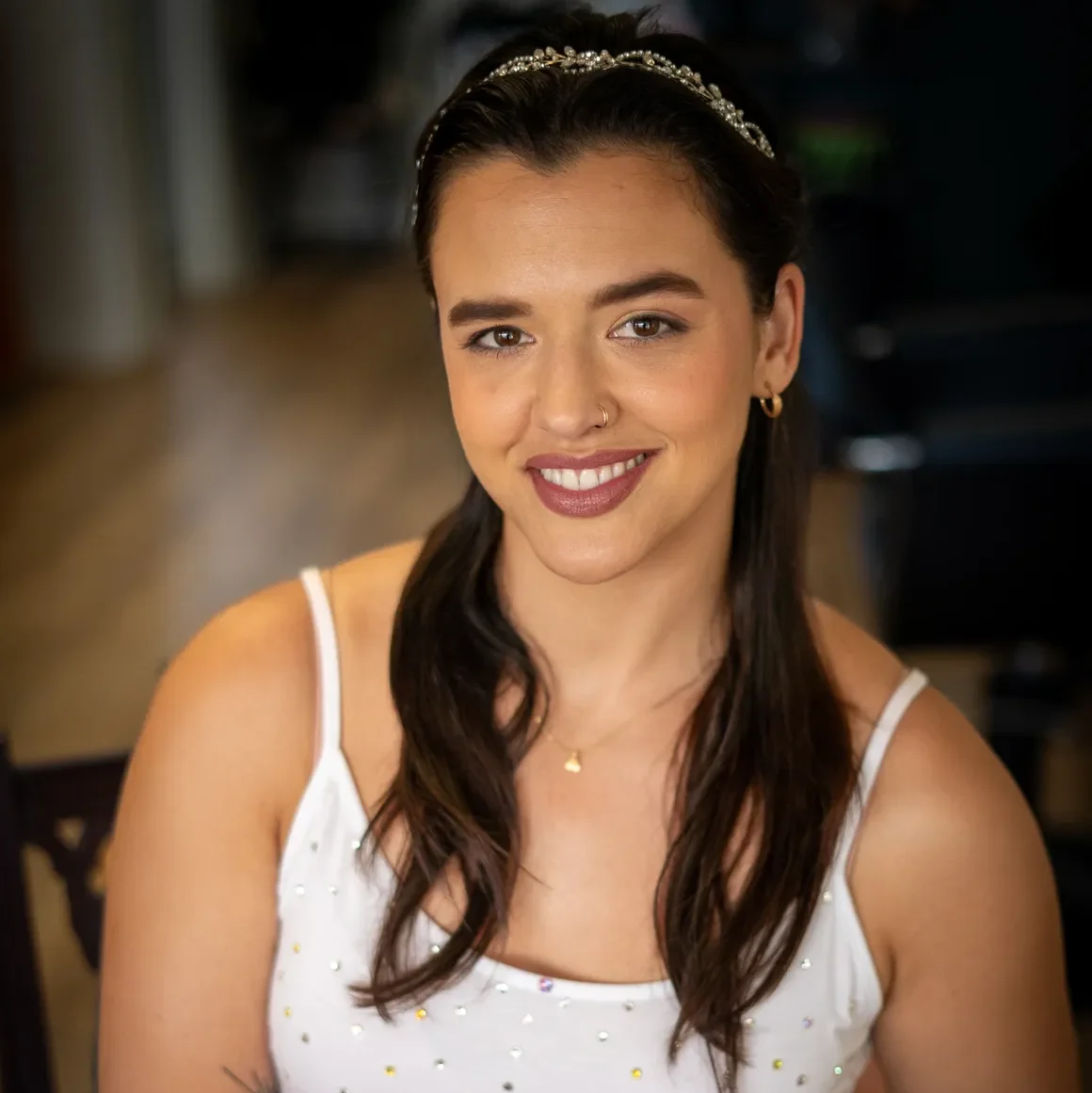 A smiling bride holds a small bouquet of pastel roses while seated indoors. She wears a white, sparkly tank top-style dress and a delicate headband, with her hair styled in soft waves. Her makeup features a warm glow, defined brows, berry-toned lipstick, and subtle shimmer eyeshadow. A tattoo is visible on her forearm, and she wears a nose ring and gold hoop earrings.